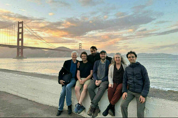 Six people sit on a low wall facing the camera with the Golden Gate Bridge and a colorful sunset in the background.