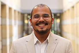 A broadly smiling man with a beard, round gold-rimmed glasses, and a silver hoop earring wears a white shirt and tan blazer, standing in a hallway.