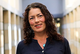 Smiling woman with dark, curly hair wearing a navy shirt and a turquoise pendant necklace in a well-lit building hallway.