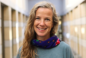 A smiling woman with long blonde and gray hair wears a light blue shirt and a dark blue floral scarf. She stands in a softly lit interior hallway.