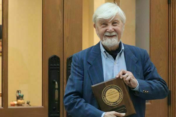 An older man with white hair and beard, wearing a blue blazer, smiles holding a wooden Founder's Medal for Lifelong Contribution plaque with a gold medallion.