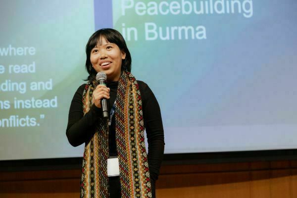 A smiling Asian woman in a black top and vibrant geometric scarf speaks into a microphone. A screen behind her displays "Peacebuilding in Burma."