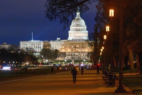 The U.S. Capitol building is brightly illuminated at night under a dark blue sky. A wide promenade with warm glowing streetlights and dark trees leads toward it, with several people walking and jogging.