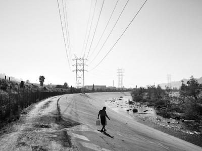 A person in a dark shirt and light shorts walks up a concrete canal bank, carrying a white bucket. Power lines and towers span the clear sky overhead, with a narrow stream flowing through the canal and sparse trees on the far bank.