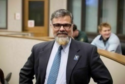 Man with dark hair and a beard, wearing a blue sports coat and blue dress shirt smiles at the camera.