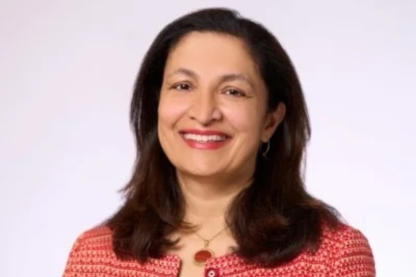 A smiling woman with dark hair wears a red patterned jacket and a gold necklace with a reddish-brown pendant.
