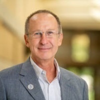 Smiling man with glasses, short hair, wearing a light blue shirt and gray blazer, in a blurred indoor academic-like setting.
