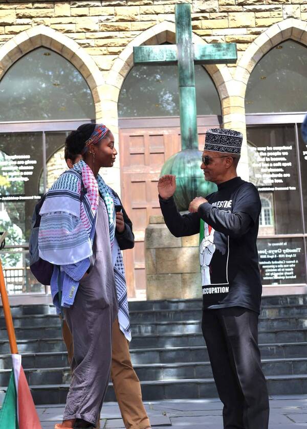 Woman in colorful headscarf and patterned keffiyeh converses with man in Kufi, black "Palestine" shirt, gesturing. Stone building, cross.