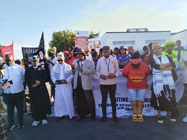 Diverse group protests. Man in beige jacket speaks into mic. Woman in black robes & colorful shawl stands. Sign: 'NO BAIL for perpetrators'.