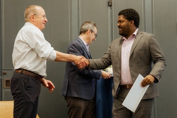 A light-skinned man in a light shirt and a dark-skinned man in a grey suit smile as they shake hands. Another man watches.