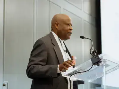 A bald, older Black man in a dark suit with a focused expression speaks at a clear podium, gesturing.