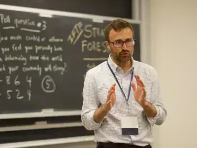 A man in a checkered shirt, glasses, and beard speaks and gestures in front of a blackboard with a poll question.