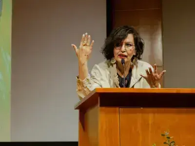 Woman with dark curly hair, glasses, and a light jacket speaks into a microphone at a wooden podium, gesturing animatedly.