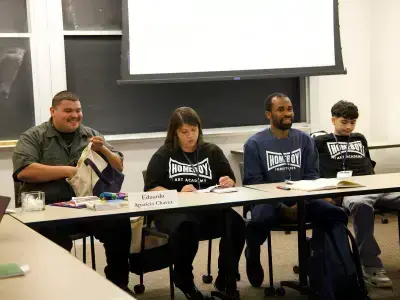 Four panelists from Homeboy Industries sit at a table. Eduardo Aparicio Chavez, wearing black, takes notes. Two men smile.