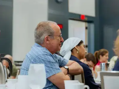 A man in a blue striped shirt and glasses smiles while seated at a table. A woman in a white head covering is behind him.