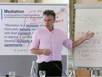 A man in a pink shirt and glasses gestures towards a whiteboard defining "Mediation" from the UN.