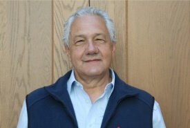A man smiles directly at the camera. He wears a white shirt, pink patterned tie, and navy blue quilted vest.