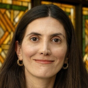 Woman with long brown hair and gold earrings smiles, with stained glass in background.