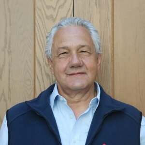 Man with graying hair smiles, wearing a blue vest over a light blue collared shirt.